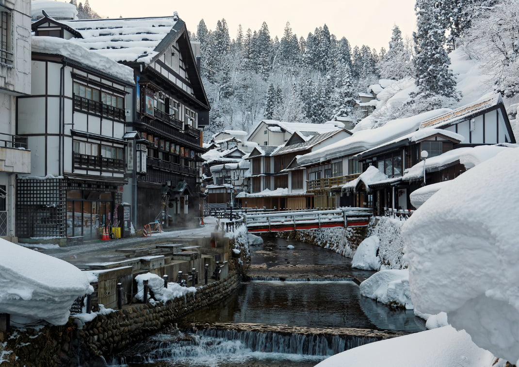 Winterlandschap in Ginzan Onsen, een beroemde Japanse hot spring bestemming in Obanazawa, Yamagata, Japan. Je ziet bruggen over een stroom met aan weerszijden historische houten gebouwen bedekt met een dikke laag sneeuw.